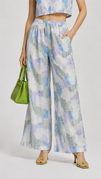 A model wearing the EVERSHE Chromaflora Ocean Lilac floral collared crop-top and wide-leg trouser co-ord set in a light blue, purple, and white watercolor print, shows the close up view of the trousers,  styled with lime green sunglasses, a green handbag, and green heels against a light gray studio background.